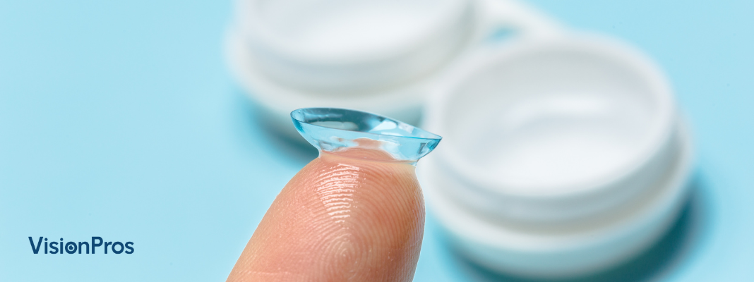 A close-up of a person’s finger holding a soft contact lens above a blue background with a white contact lens case nearby.